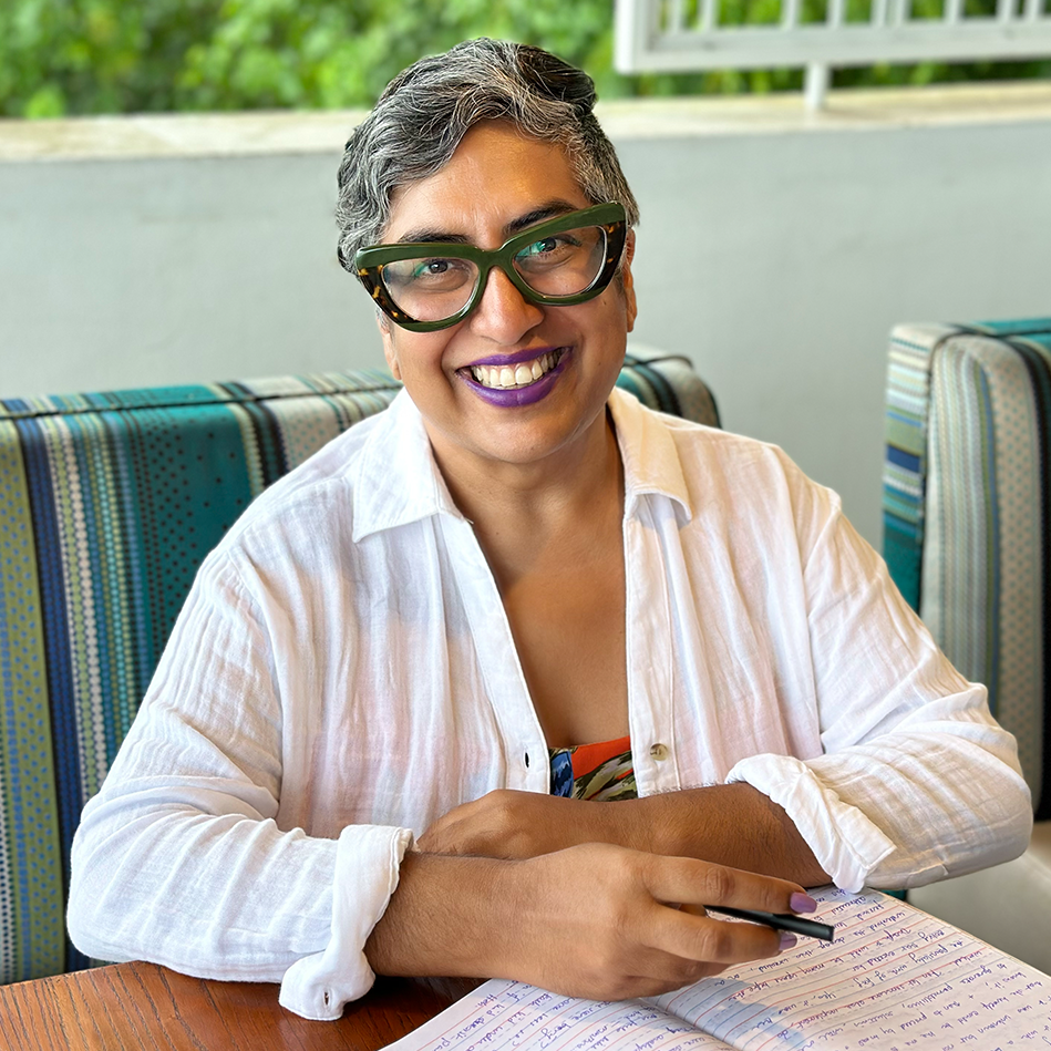 The Club’s Founder Minal Hajratwala seated on a bench at a table, pen in hand, smiling at the camera; behind her is a window open to indistinct foliage.